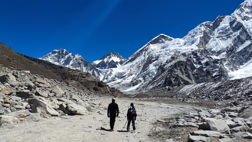 Two people walking on a rocky path towards snow-capped mountains.