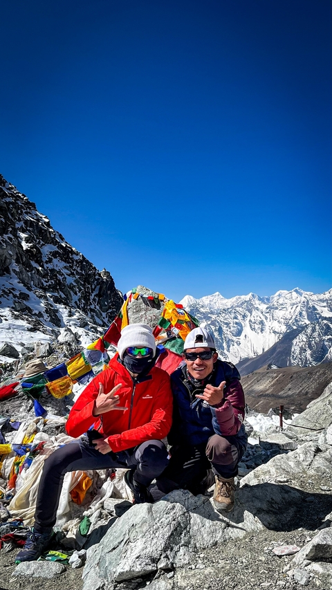 Two people in mountaineering gear with prayer flags and snow-covered mountains.