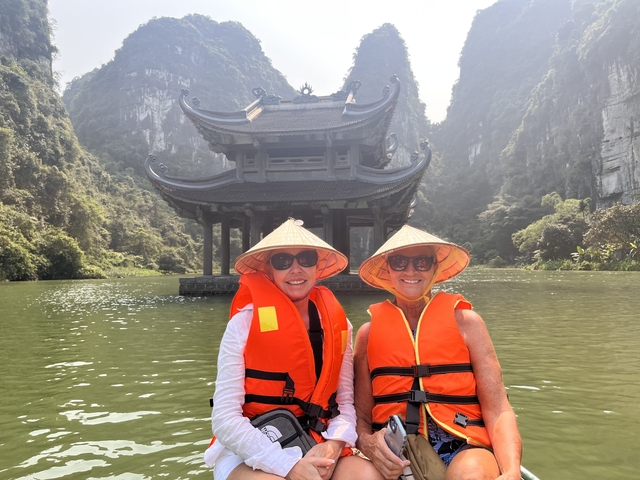 Two people in orange life vests with traditional hats in a boat in front of a pagoda.