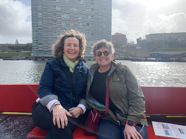       Two women sit by the waterfront with a building behind them.
  