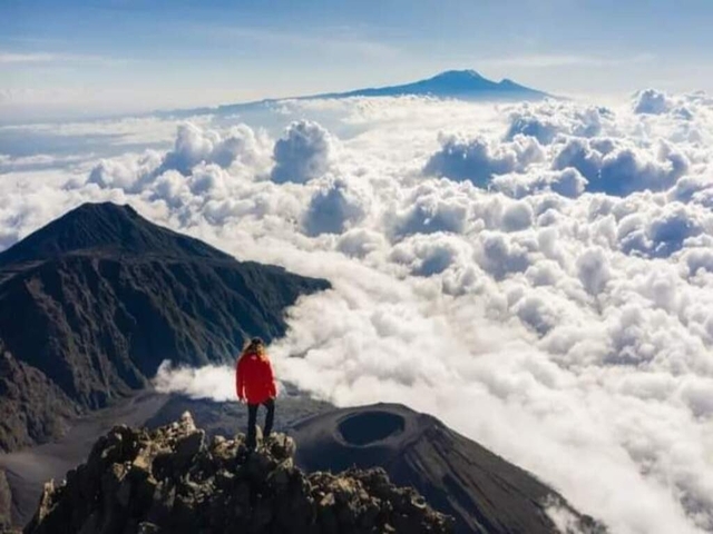 Person standing on a peak above clouds, with mountains in the distance.