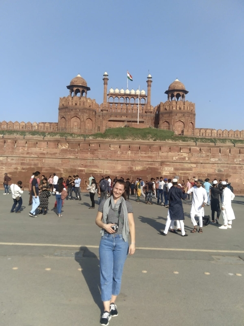 Tourist posing in front of a large red fort with people around.