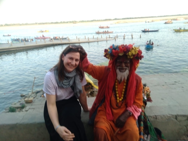       A person sitting next to a sadhu by a river with boats in the background.
  