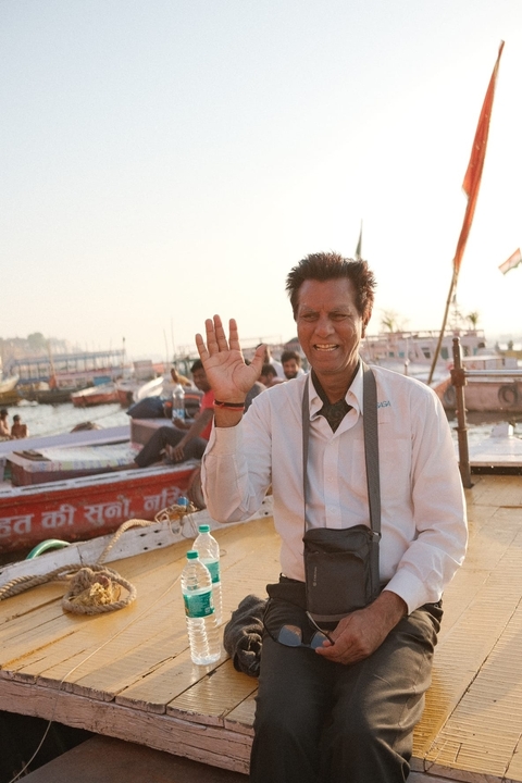       A man in a white shirt sitting on a boat, smiling and waving.
  