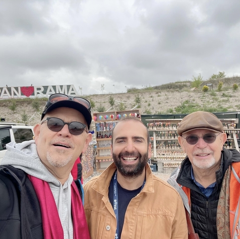 Three men taking a selfie with a souvenir stall in the background.