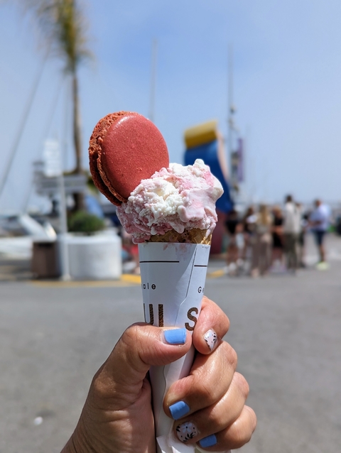       An ice cream cone topped with a macaron held up with a street view in the background.
  