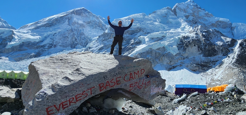       Person standing on a rock marked Everest Base Camp with snowy peaks in the background.
  