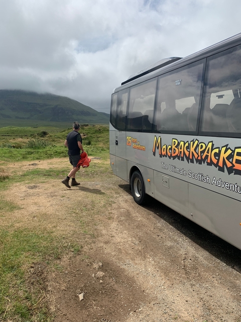 A person near a tour bus in a hilly landscape, with 'MacBackpackers' branding on the bus.