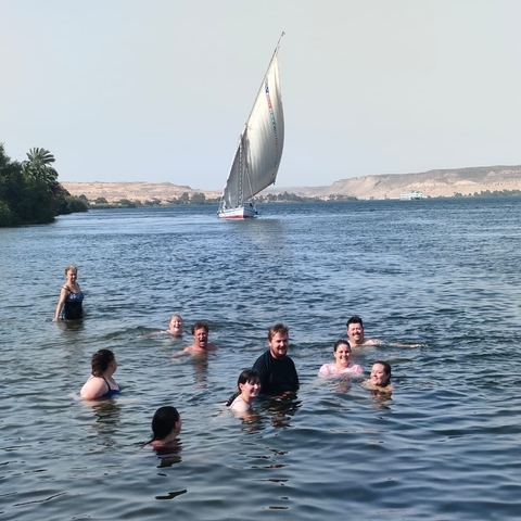 People swimming in a river with a sailboat and hills in the background.