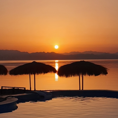       Sunset view over a calm ocean with silhouettes of distant mountains and beach umbrellas.
  
