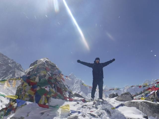 Person standing with arms raised next to a cairn with prayer flags on a snowy mountain.