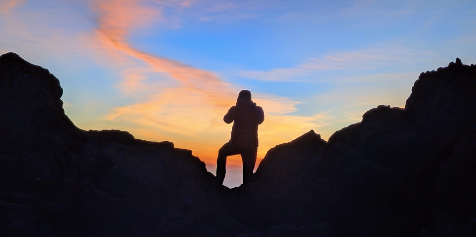 Silhouette of a person photographing the sunset from a mountain peak.