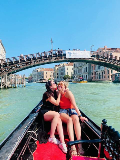 Two women on a gondola in Venice with a bridge and buildings in the background.