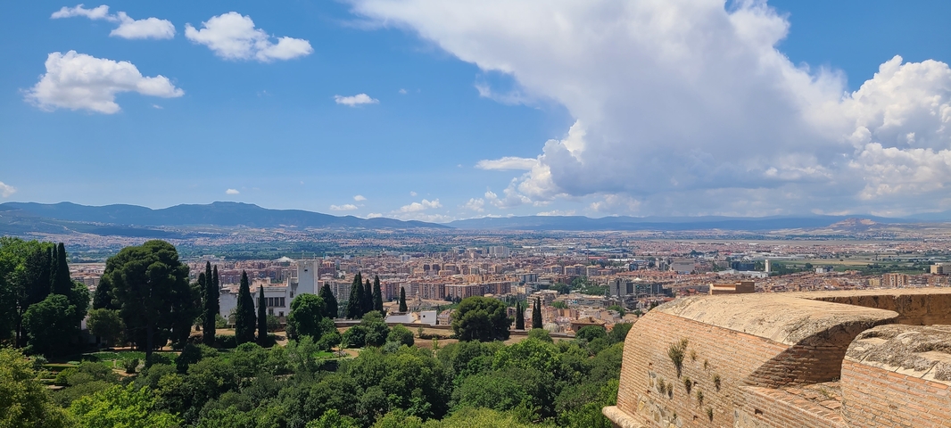       Panoramic view of a city with green landscape and distant hills.
  