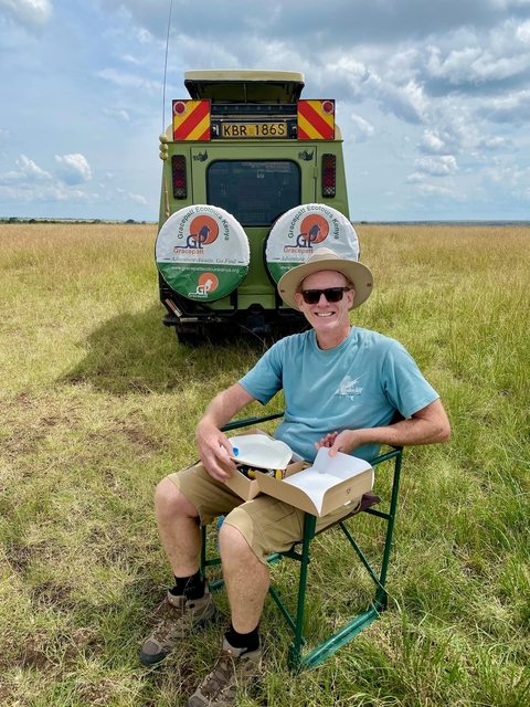       A person sitting near a vehicle in a grassy field.
  