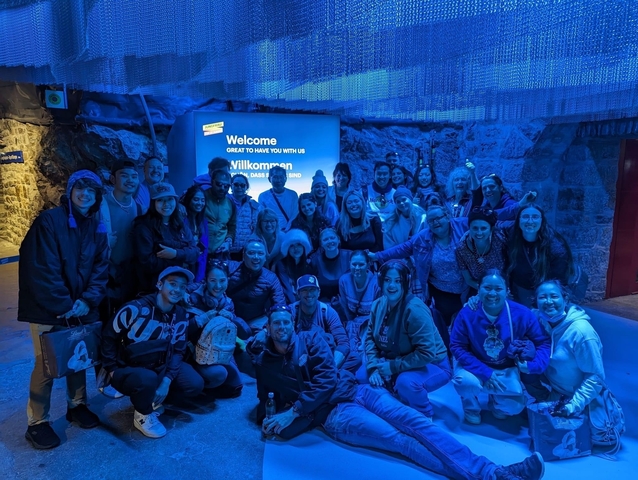 Large group posing in front of a welcome sign in a blue-lit area.