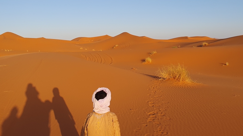       Person in traditional attire walking on desert dunes.
  