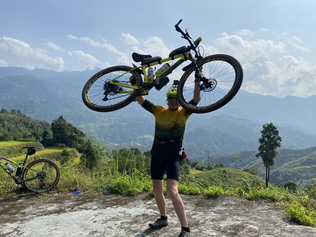       Cyclist lifting a bicycle with scenic mountains in the background.
  