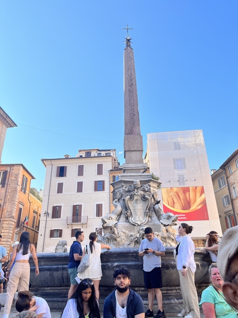       Obelisk with sculptures at its base in a square with classic buildings.
  