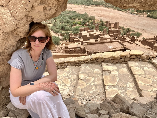 Woman sitting with a view of ancient earthen structures and green fields.