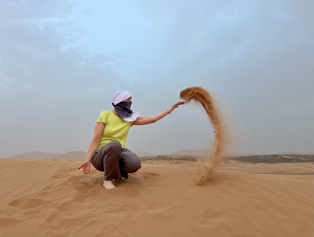 Person in desert attire playfully tossing sand in the air, set in a desert landscape.
