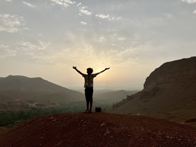       Person with raised arms standing on a hill with a dramatic sunset or sunrise.
  