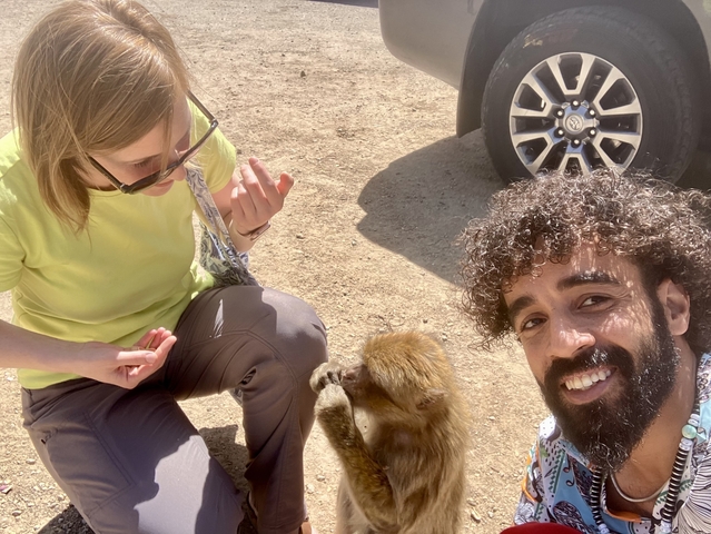       Two people interacting with a monkey, seated on the ground beside a vehicle.
  