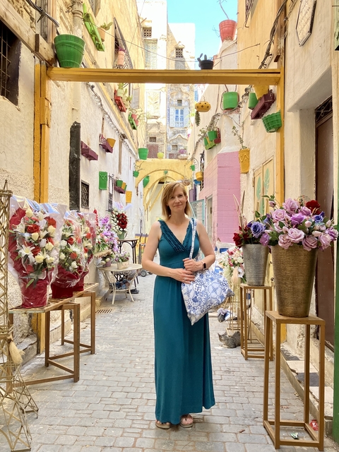       Street scene with a person posing in an alley with colorful flowers and buildings.
  