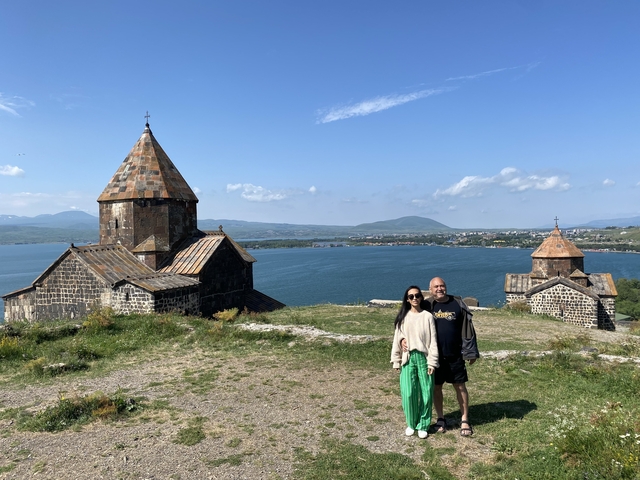 A couple standing near a lake with church buildings in the background.