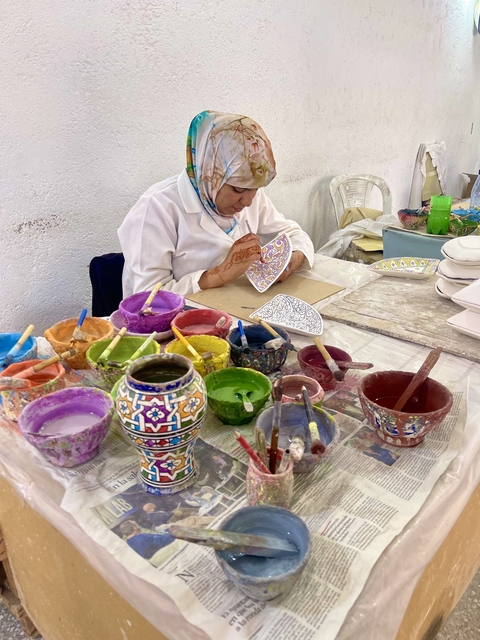 Artisan painting intricate designs on pottery with pots of colorful paints.