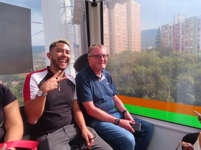       Two men smiling inside a cable car with city views outside.
  