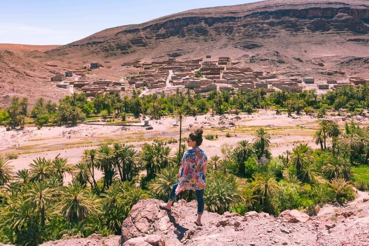       Person standing on a hill overlooking a desert village with palm trees.
  