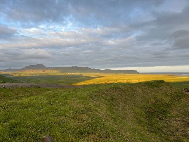       Open landscape with sunlit mountains and a green field.
  