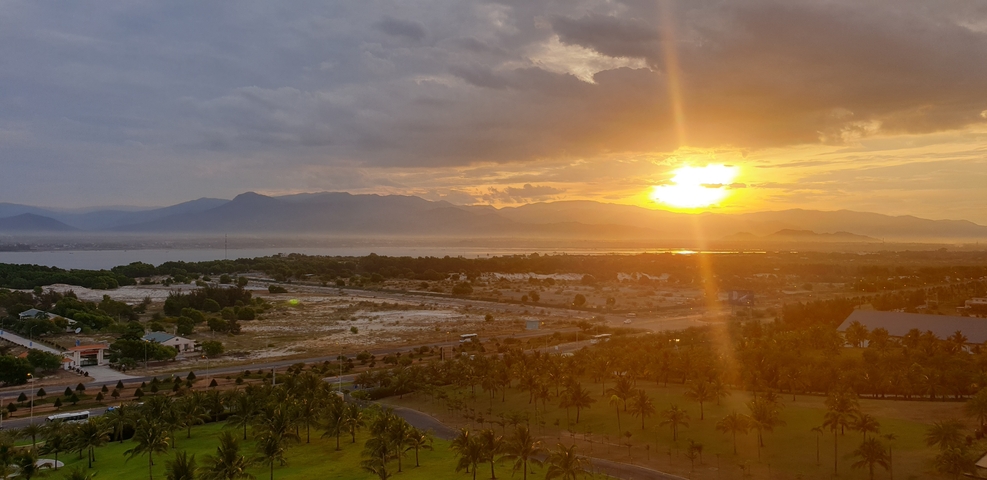 Sunset view with palm trees, mountains, and a building in the distance.