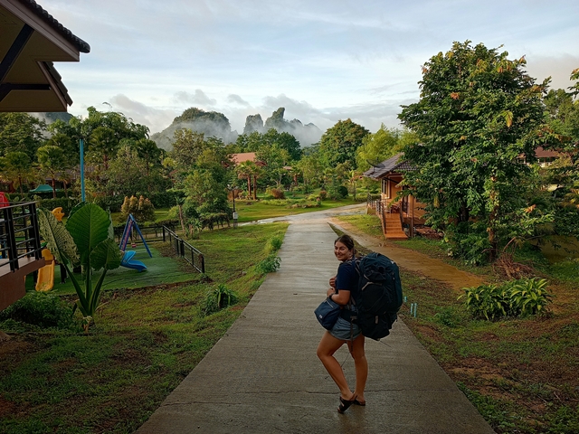 Person with a large backpack walking through a green village with mountains in the background.