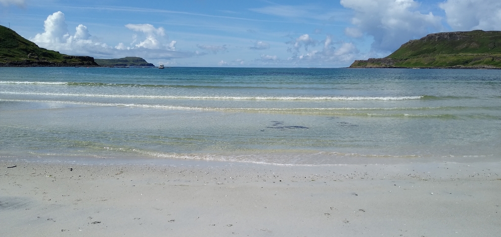 A scenic view of a sandy beach with clear blue water and distant hills.