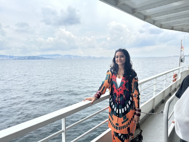       Person standing on a boat deck with a view of the sea and distant city skyline.
  