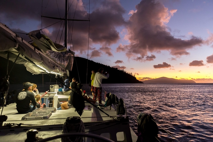 Group of people on a sailboat at sunset, observing the ocean.
