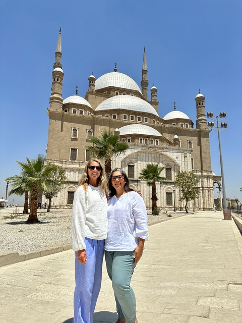       Two women pose in front of a grand mosque with palm trees.
  