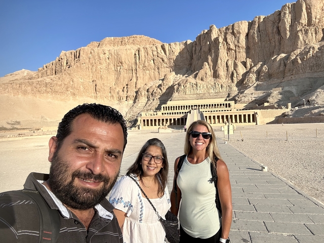       Three people standing in front of an ancient temple set into cliffs.
  