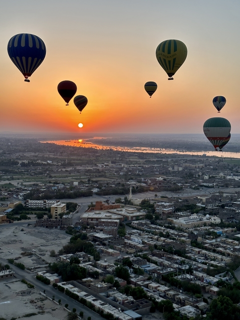       Hot air balloons floating over a city at sunset with a river in view.
  