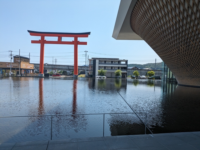       Red torii gate standing in water with modern buildings in the background.
  