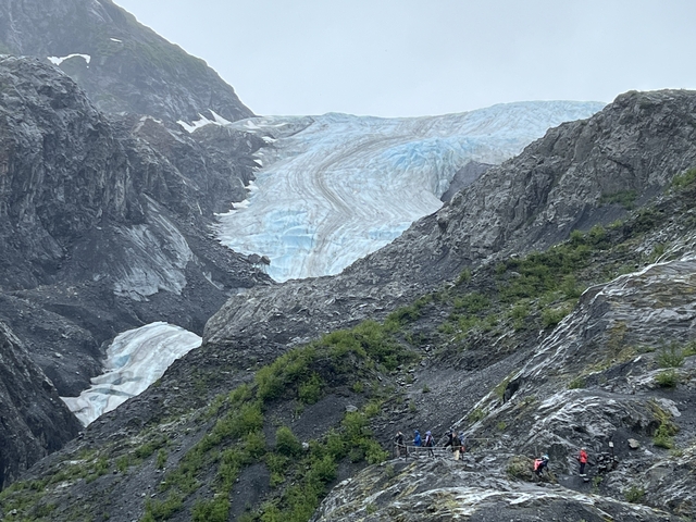       A glacier with hikers visible in the rocky foreground.
  