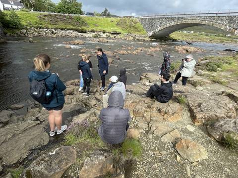       People sitting and standing on rocks by a river under a stone bridge.
  