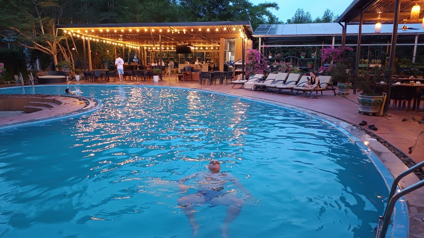 A man floating in a pool near a lively restaurant area.