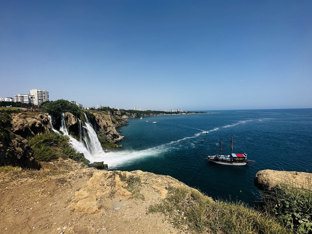       Waterfall by the sea with a sailing ship nearby.
  