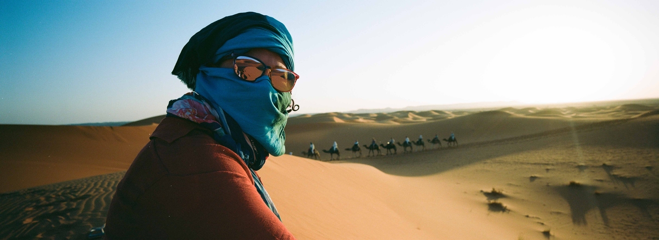       Person wearing a scarf in the desert with camels in the background.
  