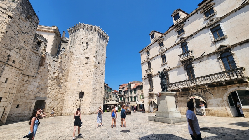       People walking in a historical square with ancient stone towers.
  