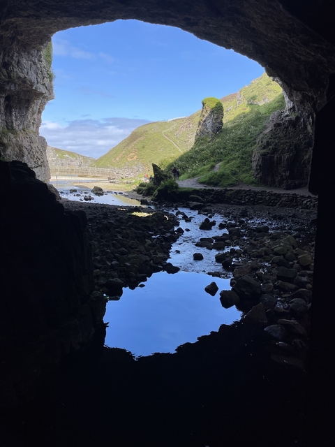 A cave entrance with a view of a rocky path and a wooden bridge in the background.