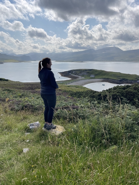       A person standing on a hilltop overlooking a scenic bay and distant hills.
  